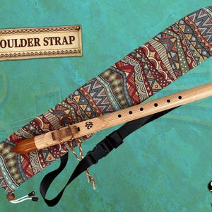 May include: A wooden flute with a brown mouthpiece and a patterned case. The case has a black shoulder strap and the text "SHOULDER STRAP" on a sign. The flute is light brown with several finger holes.