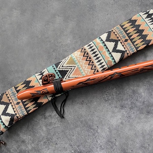 Può includere: Un flauto di legno con intagli intricati, riposante in una custodia di tessuto con motivo colorato. La custodia ha una chiusura con coulisse e un fiocco.