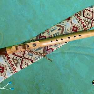 May include: A wooden Native American flute with a brown leather mouthpiece and a decorative patterned fabric carrying case. The flute has six holes and is resting on top of the case.
