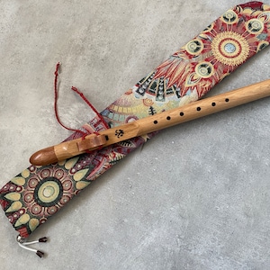 Peut inclure: Une flûte en bois brun clair avec six trous et un bloc sculpté, posée sur un étui en tissu long orné d'un motif circulaire vibrant rouge, doré et noir. La flûte présente un petit symbole noir imprimé sur sa surface et un cordon rouge.