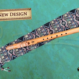 May include: A wooden flute with a leather strap and a case with a geometric pattern. The flute has six holes and is accompanied by a case with a black, blue, and pink design. The words "NEW DESIGN" are displayed on a banner.