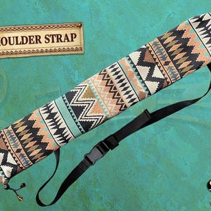 May include: A patterned shoulder strap case in black, beige, teal, and orange, with a black adjustable strap and buckle. The case has a cylindrical shape. The text "SHOULDER STRAP" is displayed on a wooden sign.