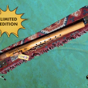 May include: A wooden flute with finger holes is displayed in a patterned case with a fringed edge. The case is red, brown, and cream-coloured. A tag hangs from the flute. The words "LIMITED EDITION" are in a yellow burst shape.