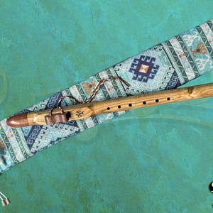 Può includere: Un flauto di legno con bocchino marrone e dettagli in pelle è appoggiato su una custodia blu e bianca fantasia. Il flauto ha diversi fori per le dita e un cinturino decorativo in pelle. La custodia presenta un design geometrico. Lo sfondo è verde acqua.