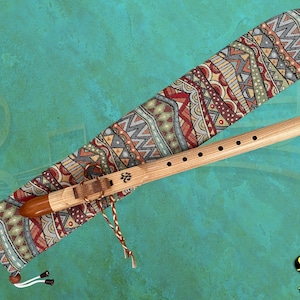 Könnte beinhalten: Eine indianische Holzflöte mit sechs Löchern, die in einem farbenfrohen, gemusterten Stoffetui liegt. Das Etui hat einen Kordelverschluss und einen Quasten. Die Flöte ist aus Naturholz gefertigt und hat ein schlichtes Design. Das Etui ist mit geometrischen Formen und Mustern in Rot-, Orange-, Gelb-, Grün- und Blautönen verziert.