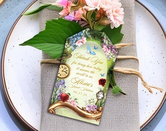 May include: A light blue and white place setting with a gray napkin and a floral arrangement. A thank you tag with a whimsical Alice in Wonderland theme is tied to the napkin. The tag reads "Thank You for being here today at what is to be Alice's Wonderland Tea Party".