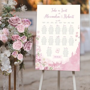 May include: A pink and white wedding seating chart with a castle silhouette and floral accents. The chart lists the names of guests assigned to each table, numbered one through twelve. The text at the top reads "Take a Seat with Alexandra & Robert".