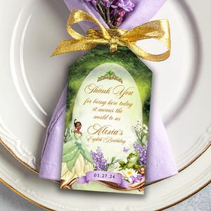 May include: A place setting with a white plate, gold rim, and a lavender napkin. A gold fork and a glass of purple liquid are on the table. A gift tag with text and a gold bow is attached to the napkin. Lilac flowers are arranged around the plate.