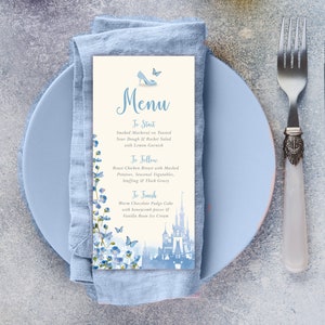 Może przedstawiać: Jasnoniebieska karta menu z motywem Kopciuszka. Menu zawiera szklany pantofelek, motyle i akwarelowe przedstawienie zamku Kopciuszka. Menu zawiera trzydaniowy posiłek: Wędzony makrela, Pieczeń z kurczaka i Ciepły tort czekoladowy z polewą.