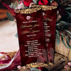 Op de afbeelding: Twee bordeauxrode huwelijksprogramma's met gouden sierlijke frames en tekst. De programma's luiden "Welkom bij de bruiloft van Alexandra Jones en Robert Spencer 31 augustus 2026." Bloemschikkingen van rode rozen en groen omringen de programma's.