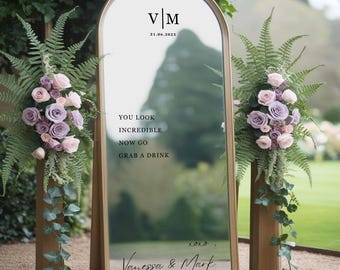 Puede incluir: Un espejo arqueado con marco dorado, con las iniciales "V|M" y la fecha "21.06.2025". El espejo tiene el texto "YOU LOOK INCREDIBLE NOW GO GRAB A DRINK" y los nombres "Vanessa & Mark". Arreglos florales a ambos lados.