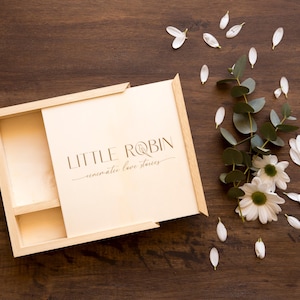 May include: A wooden box with a hinged lid, open to reveal a compartment with the text "LITTLE ROBIN -  memorable love stories". The box is on a brown wooden surface with white flower petals scattered around it.