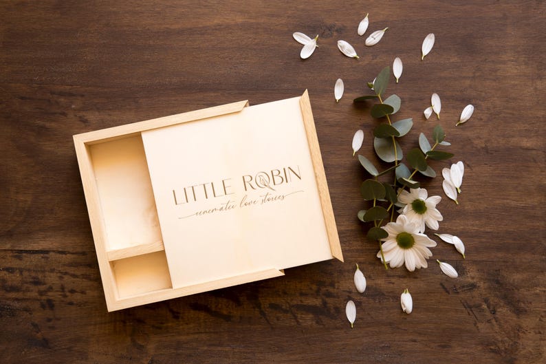 May include: A wooden box with a hinged lid, open to reveal a compartment with the text "LITTLE ROBIN -  memorable love stories". The box is on a brown wooden surface with white flower petals scattered around it.