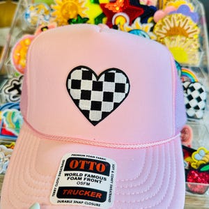 Op de afbeelding: Een lichtroze truckerpet met een zwart-wit geblokt hartontwerp. De pet heeft een mesh achterkant en een gebogen klep. De pet heeft de tekst "OTTO WORLD FAMOUS FOAM FRONT OSFM TRUCKER" op een label.