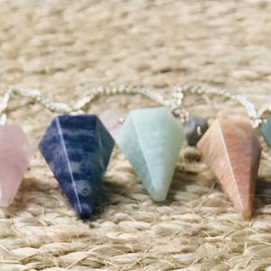 May include: Six crystal pendulums of various colors, including pink, blue, green, and brown, are strung together on a silver chain. The pendulums are all in the shape of a pyramid.