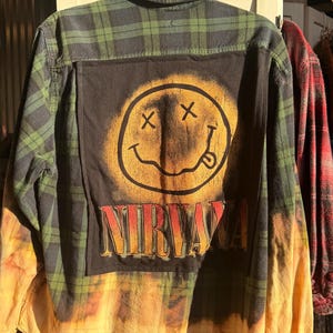 Puede incluir: Una camisa de manga larga con botones, patrón de cuadros verdes y negros y una sección inferior amarilla desteñida. Un gran parche cuadrado negro en la espalda muestra una cara sonriente amarilla pintada con aerosol y la palabra NIRVANA en letras rojas.
