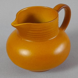 May include: A brown ceramic pitcher with a rounded body and a handle. The pitcher has a smooth, glossy finish.