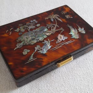 May include: A rectangular box with a dark brown tortoiseshell-patterned finish featuring an iridescent mother-of-pearl inlay depicting a coastal landscape with a house, palm trees, and a boat. The box has a gold-toned metal latch on the front.