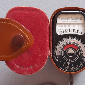 May include: A brown leather case with a red interior is open to reveal a black Weston Master III Universal Exposure Meter. The meter has a dial with numbers from 25 to 1600 and a needle that points to 100. The meter also has a rotating dial with numbers from 1 to 100.
