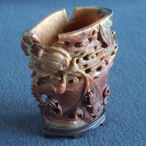 May include: A carved stone vase with detailed relief carvings. The vase depicts a dragon and fish design, in tan and reddish-brown tones. The top is curved, and the vase rests on a dark wooden base. The height is approximately 15 cm.