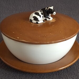 May include: A white ceramic butter dish with a brown wooden lid and a black and white cow figurine on top.