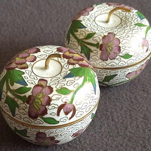 May include: Two apple-shaped decorative boxes with lids. The boxes are white with a floral design in pink, green, and gold. Each box has a gold-colored stem. The boxes are on a gray surface.