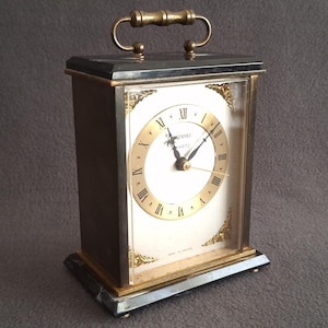 May include: A rectangular quartz clock with a gold-tone face and Roman numerals. The clock has a dark brown wooden frame, a gold handle, and decorative gold accents. The clock face reads "STAMMEL QUARTZ".