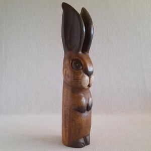 May include: A tall, upright wooden rabbit figurine carved from a single piece of wood. The sculpture features a dark brown finish on the long, pointed ears and base, with a lighter brown body, a carved snout, and visible wood grain throughout.