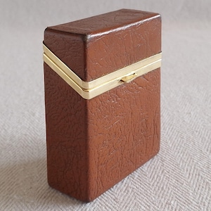 May include: A rectangular brown leather case with a textured surface. The case has a gold-colored metal trim around the edges and a small clasp. The case is closed and appears to be a container for small items.