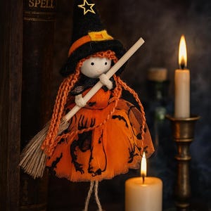 May include: A handcrafted Halloween witch doll with orange hair, a black hat with an orange band, and an orange dress. The doll holds a broom and is suspended by a string. The background includes candles and a vintage book.