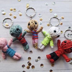 May include: Five handmade yarn doll keyrings in various colours: pink, teal, tan, white, and red. Each doll has a round head, button eyes, and is wrapped in yarn. The dolls are lying on a white wooden surface with scattered gold and silver sequins.