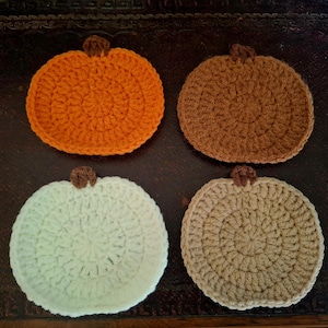 May include: Four handmade crochet pumpkin coasters in orange, brown, white, and tan. Each coaster has a circular design with a small brown stem at the top. The coasters are arranged on a dark brown surface.