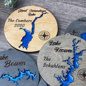 May include: Four wooden ornaments with blue epoxy resin lake outlines and compass designs. The ornaments are personalized with names and dates, including "The Combers 2020", "Eberhard's 2020", "The Schuhlens", and "The Spooner Family".