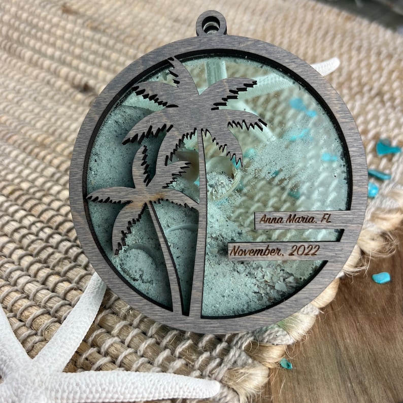 May include: A round wooden ornament with a cutout of two palm trees and a clear resin insert containing sand and seashells. The ornament is inscribed with "Anna Maria, FL" and "November, 2022".