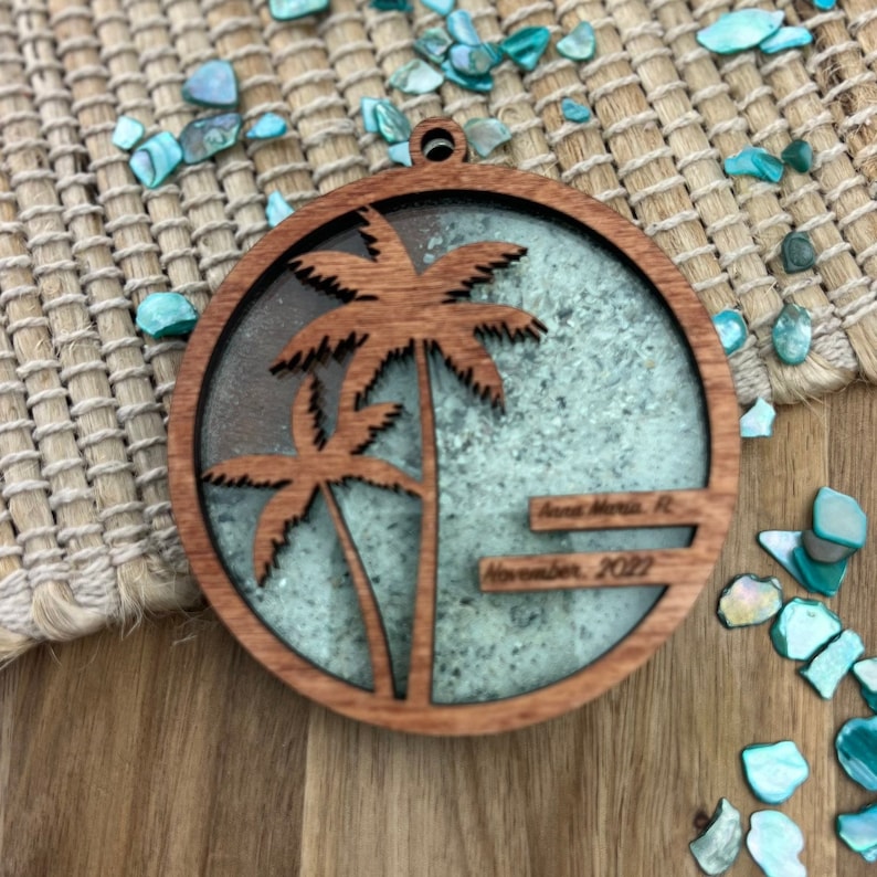May include: A round wooden ornament with a cutout of two palm trees. The ornament is filled with blue and green glitter and has the text "Anna Maria, FL" and "November, 2022" written on it.
