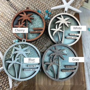May include: Four round wooden ornaments with palm tree silhouettes and sand inside. The ornaments are in the colors cherry, white, blue, and gray. Each ornament has a small tag with the location and date: Siesta Key, FL November 2022, Anna Maria, FL November 2022, Siesta Key, FL November 2022, and Siesta Key, FL November 2022.