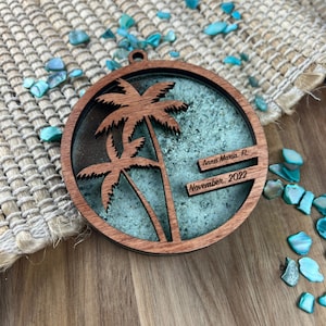 May include: A round wooden ornament with a teal blue background and two cutout palm trees. The ornament has the text "Anna Maria, FL" and "November, 2022" engraved on it.