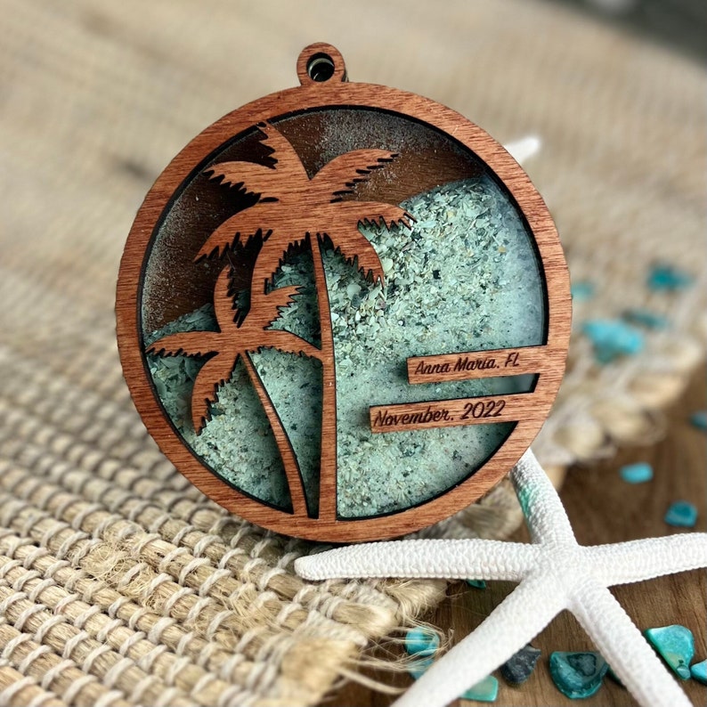 May include: A round wooden ornament with a cutout of two palm trees and a layer of green sand inside. The ornament has the text "Anna Maria, FL" and "November, 2022" inscribed on it.