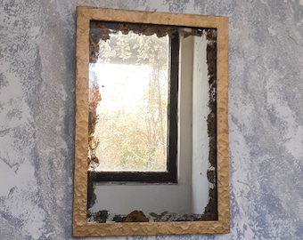 May include: A gold-framed mirror with a distressed finish hangs on a textured wall. The mirror reflects a window with a view of trees outside.