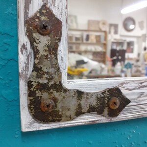 May include: A whitewashed wooden frame with a distressed finish, featuring a metal corner brace with visible rust and screws. The frame is against a teal background.