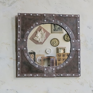 May include: A round mirror with a rustic, industrial-style frame. The frame is made of metal and has a dark brown finish with silver accents. The mirror is surrounded by a decorative border of rivets.