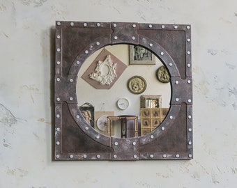 May include: A round mirror with a rustic, industrial-style frame. The frame is made of metal and has a dark brown finish with silver accents. The mirror is surrounded by a decorative border of rivets.