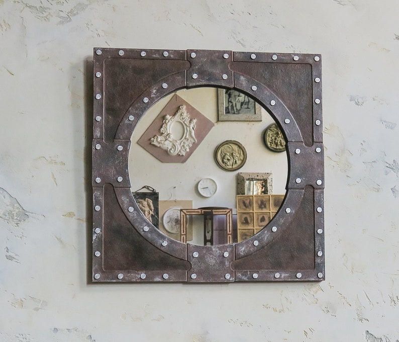 May include: A round mirror with a rustic, industrial-style frame. The frame is made of metal and has a dark brown finish with silver accents. The mirror is surrounded by a decorative border of rivets.