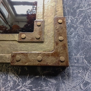 May include: Close-up of a corner of a mirror with a rustic, distressed metal frame. The frame is decorated with brass rivets and has a brown and gold finish.