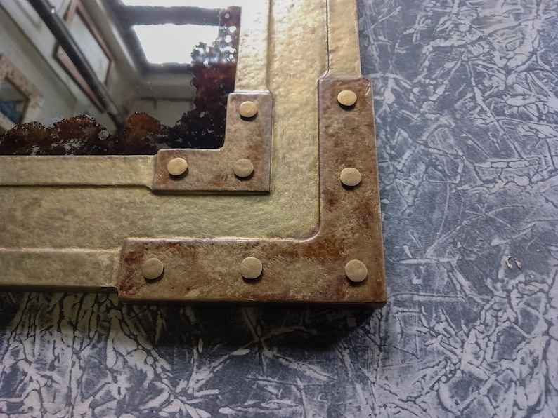 May include: Close-up of a corner of a mirror with a rustic, distressed metal frame. The frame is decorated with brass rivets and has a brown and gold finish.