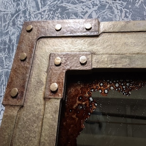 May include: A close-up of a corner of a rustic mirror with a gold and brown frame. The frame has a layered design with rivets and a distressed finish. The mirror is reflecting a blurry image.