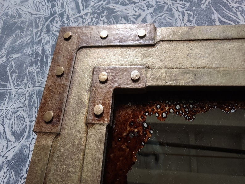 May include: A close-up of a corner of a rustic mirror with a gold and brown frame. The frame has a layered design with rivets and a distressed finish. The mirror is reflecting a blurry image.