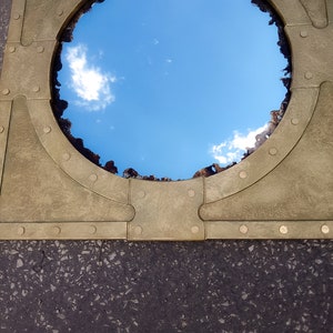 May include: A round mirror with a brass frame and rivets. The mirror reflects a blue sky with white clouds.