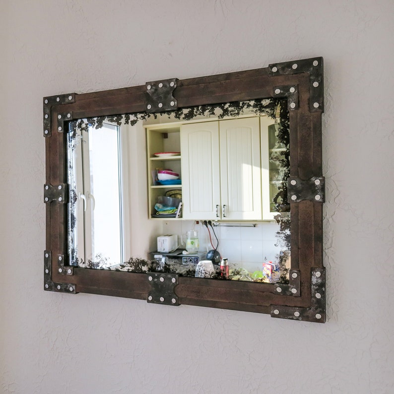 May include: A brown wooden framed mirror with metal accents. The mirror reflects a kitchen with white cabinets and a window.