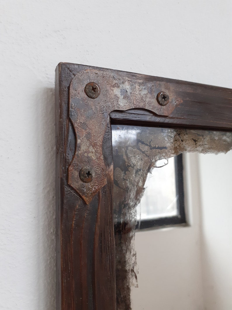 May include: A rustic wooden mirror with a distressed, brown finish. The mirror has a metal frame with three visible screws. The frame is decorated with a rusty patina.